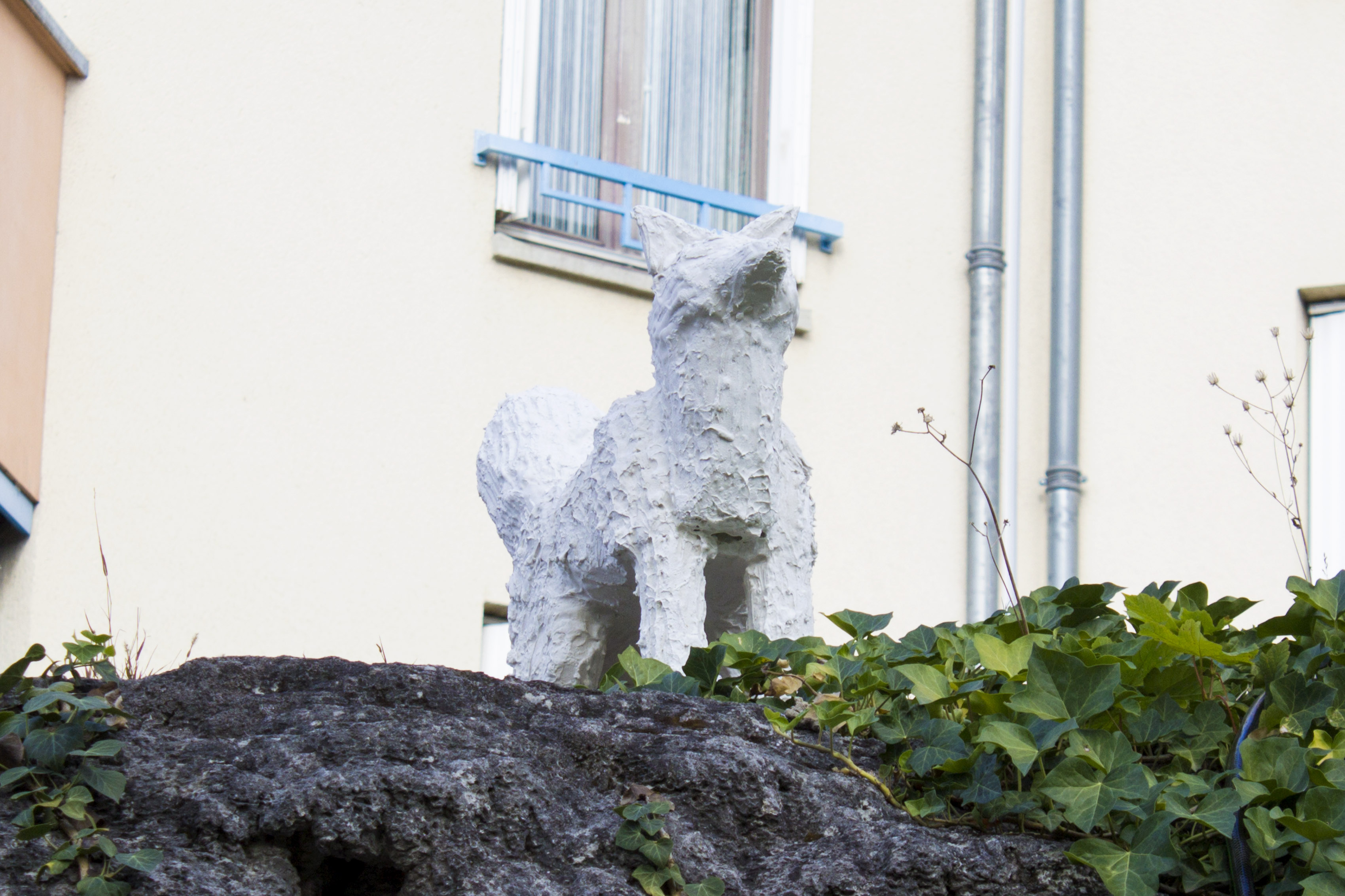 Sculture d'un renard adlte et debout en plâtre blanc effet crépi, posé sur un talus.